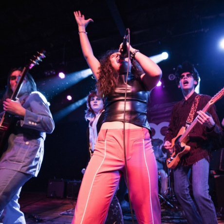 A young woman singing into a microphone while surrounded by musicians playing instrments